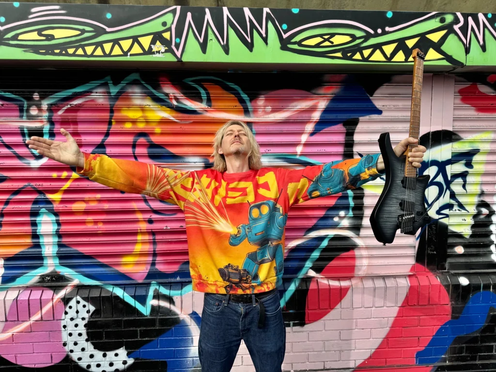 Tim Hutch holding his Strandberg guitar in front of colourful street art